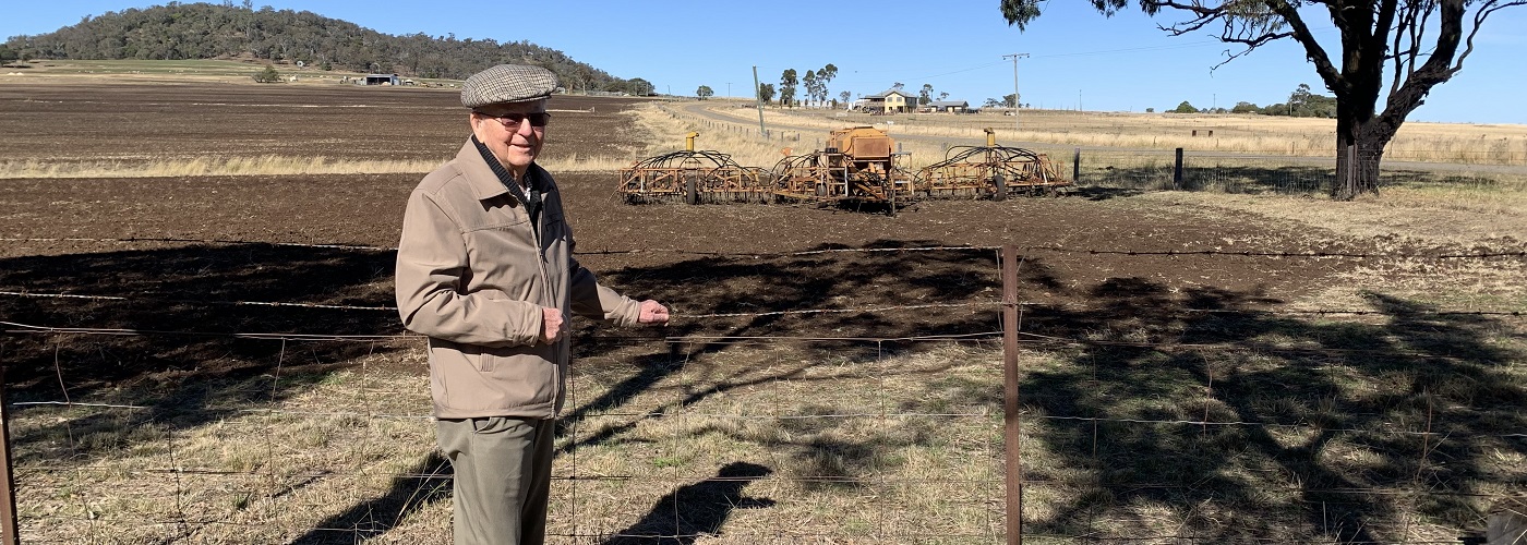 An elderly gentleman stands in front of a paddock where a seeder can be seen in the background