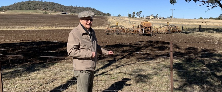 An elderly gentleman stands in front of a paddock where a seeder can be seen in the background An elderly gentleman stands in front of a paddock where a seeder can be seen in the background